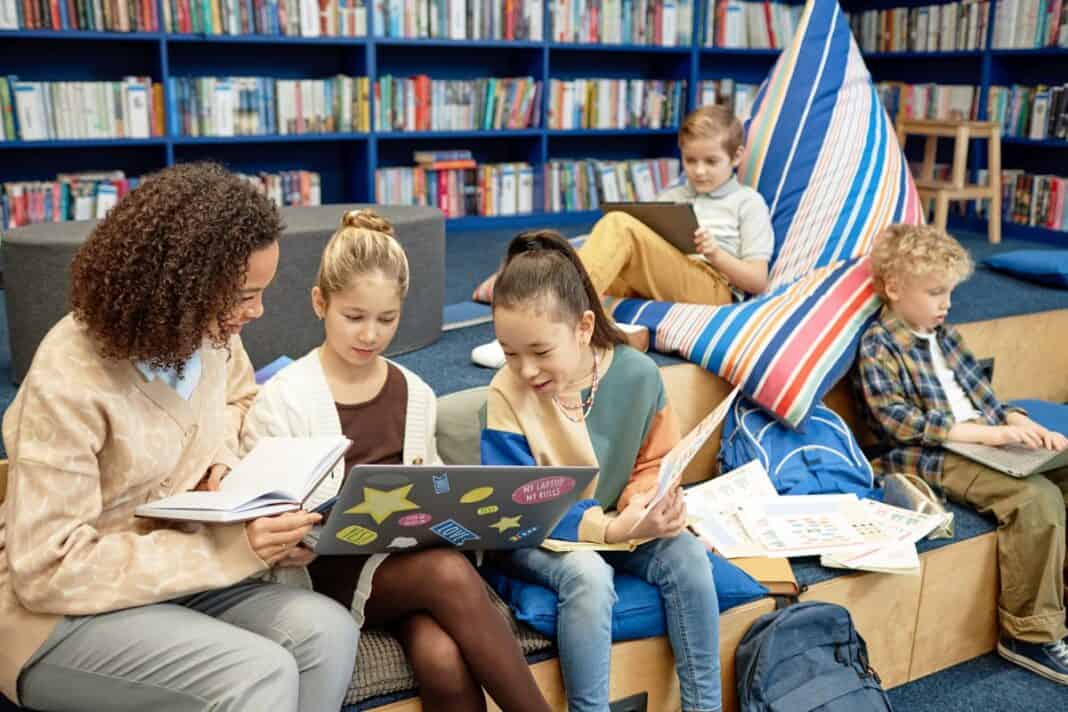 Two Girls using Laptop in Library with Teacher Helping Buchclub der Kinder
